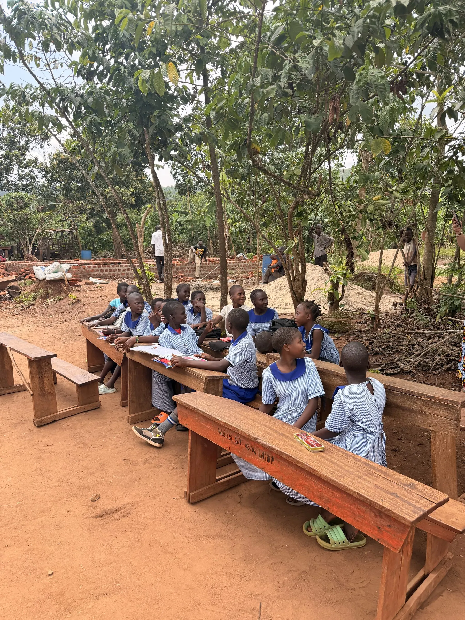Students in uniform at wooden desks, holding class outdoors beneath the trees